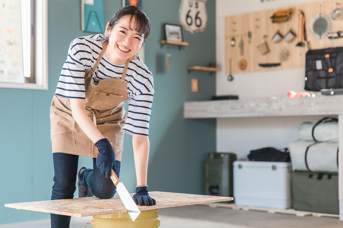 家のガレージでノコギリで木を切るアジア人女性(DIY教室・ワークショップ・日曜大工・体験教室)
